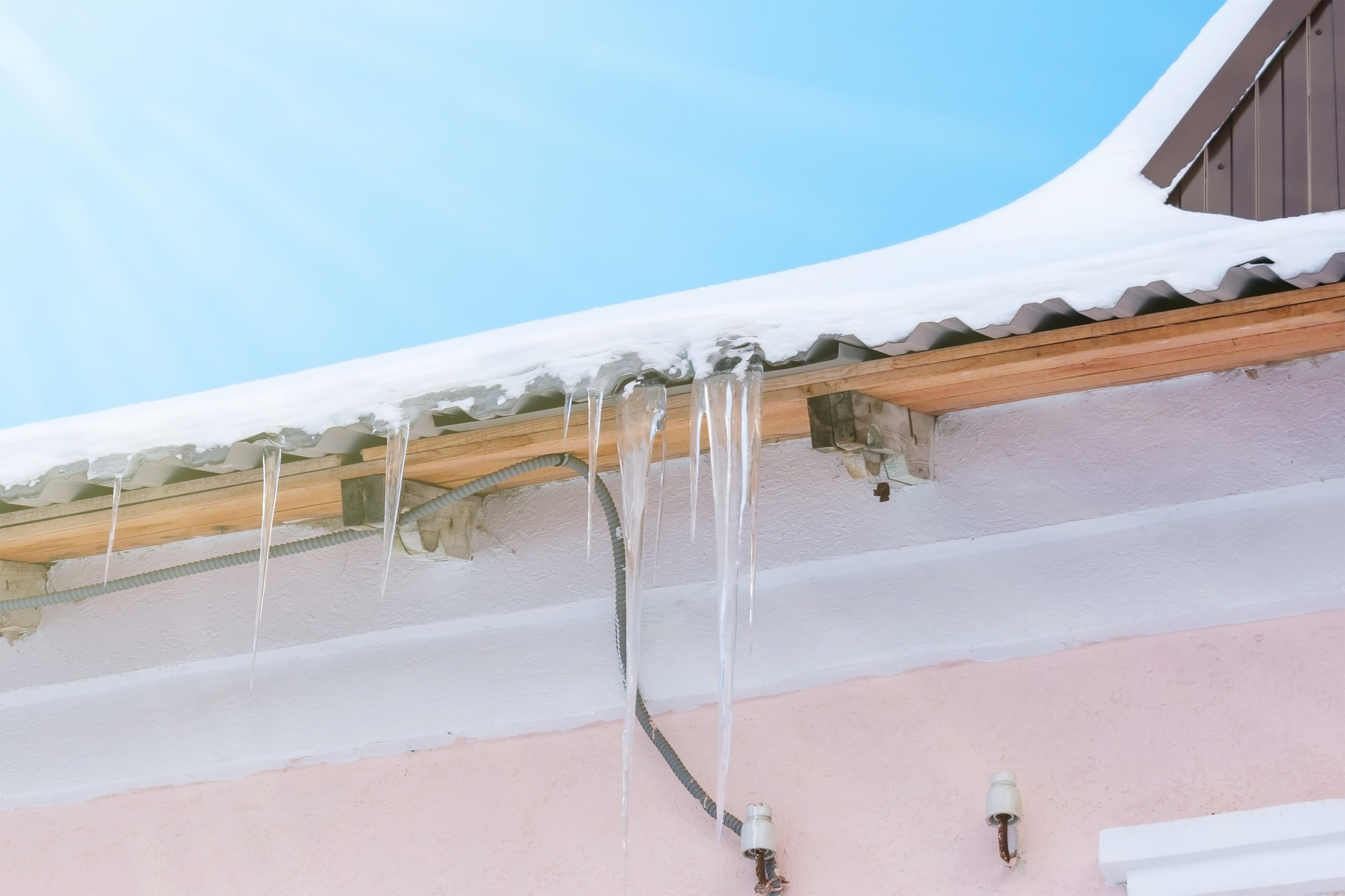 A close-up of a house roof covered in snow with several hanging icicles highlights common roofing issues. The sky is clear and bright blue, with sunlight creating a crisp winter atmosphere. The house is light pink with visible exterior wiring and light fixtures.