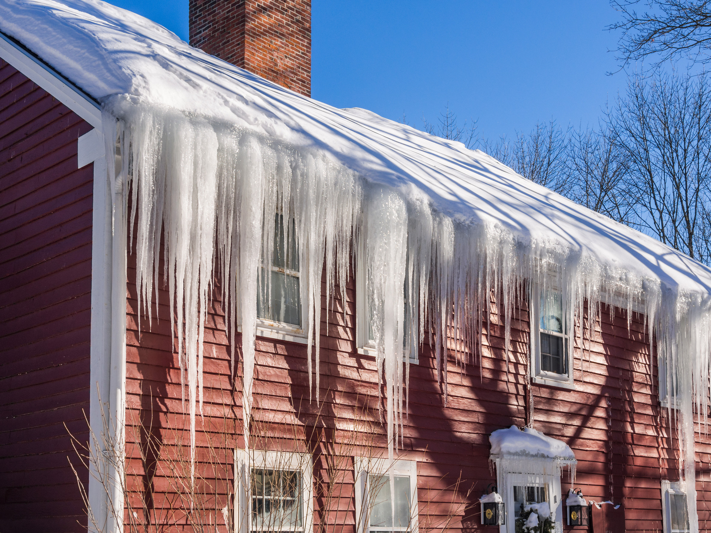 A red wooden house with a snow-covered roof adorned with large icicles hanging down. The bright blue sky and bare trees in the background create a wintry scene.