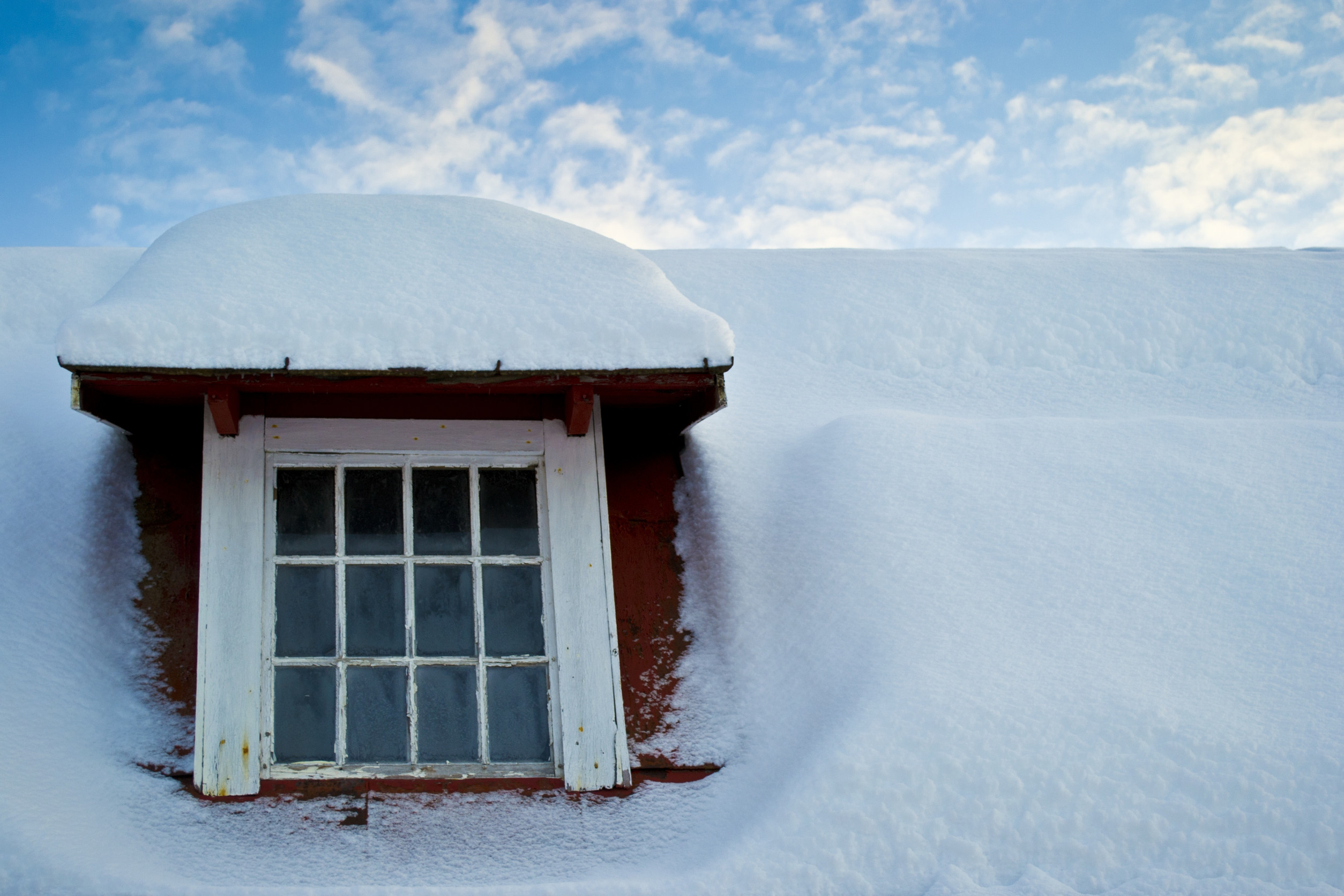 A snow-covered roof with a small window partially buried in snow, designed to protect against the harsh winter weather, is framed against a blue sky with scattered clouds.