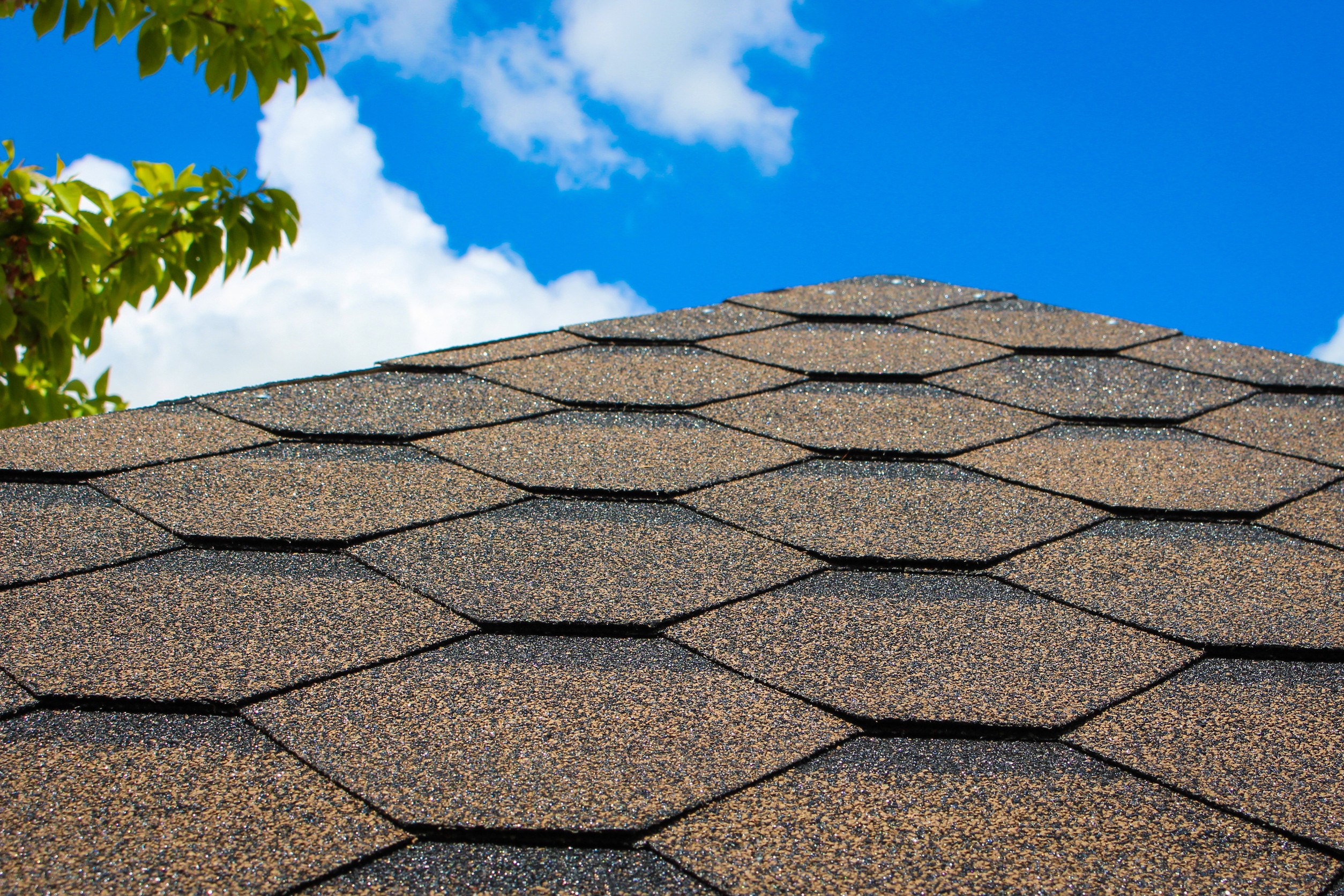 Close-up of a brown shingle roof under a bright blue sky with a few clouds—a testament to choosing the best roofing material. A small section of a leafy green tree is visible in the upper left corner, adding a touch of nature to this architectural scene.