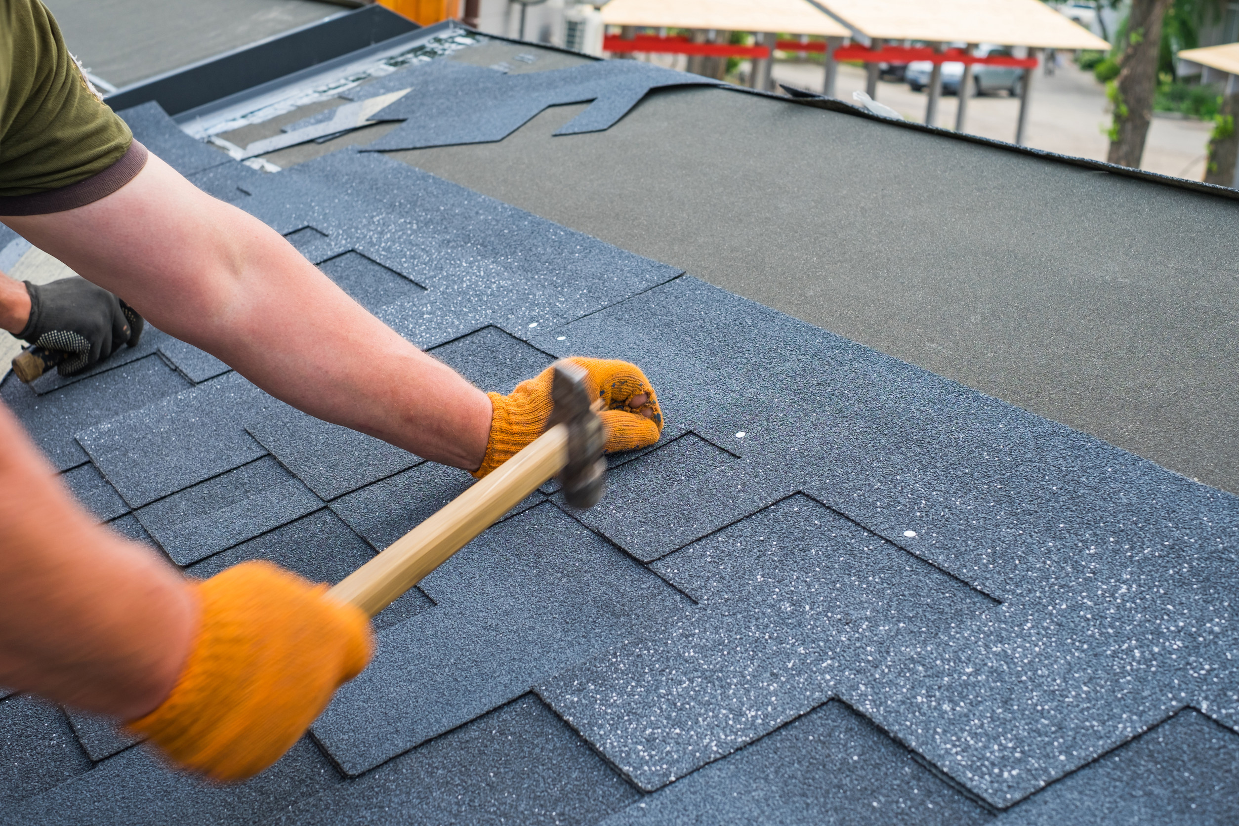 A person wearing gloves is using a hammer to nail down roofing shingles, expertly ensuring that replacing your roof offers maximum benefits. The dark gray shingles contrast against the background of other visible roofing materials, with hands intently focused on the task at hand.