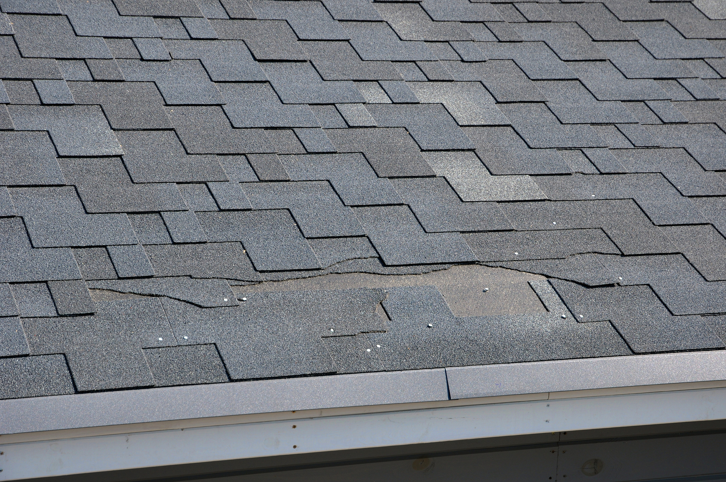 A close-up view of an asphalt shingle roof shows visible damage, with cracked and slightly lifted shingles revealing a potential leak hazard. This could indicate it's time to replace your roof. The edge of the roof and the gutter are visible at the bottom.