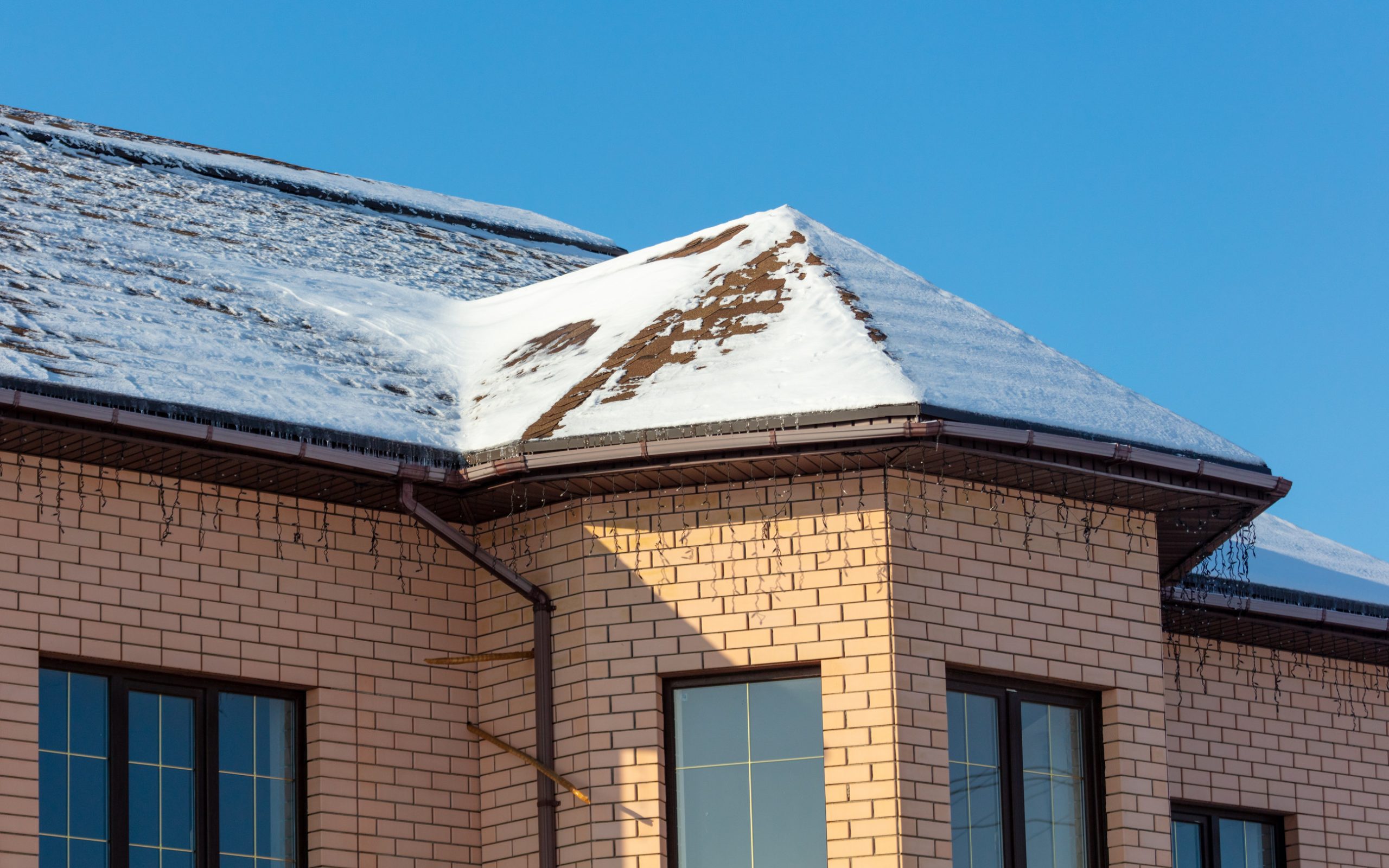 A brick building corner with a sloped roof partially covered in snow. Snow is melting, creating icicles hanging from the eaves. The sky is clear and blue, casting a shadow on the building.