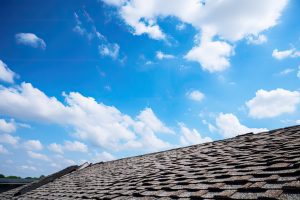 A rooftop with gray shingles is shown in the foreground, slanting upwards—a reminder for homeowners to add roof maintenance to their checklist. The bright blue sky above is dotted with fluffy white clouds on a sunny day.