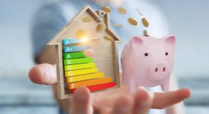 A hand holds a wooden home energy efficiency chart shaped like a house, labeled from A++ to F, next to a pink piggy bank with gold coins dropping in, symbolizing home energy savings and cost efficiency.