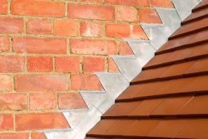 Close-up of a brick wall with step-shaped metal flashing meeting a sloped, brown tiled roof, showing the intersection detail between the wall and roof for weatherproofing.