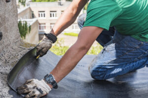 A person wearing gloves, a green shirt, and a watch is kneeling and installing a sheet of roofing material on a flat roof. Buildings are visible in the blurred background.