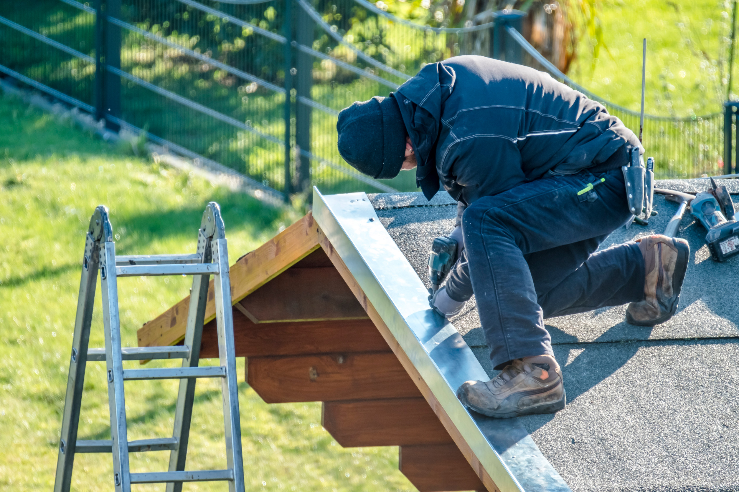 A worker wearing a jacket and beanie kneels on a roof, using a power tool to secure metal flashing along the edge. A ladder leans against the roof, and tools are scattered nearby. The background shows grass and a fence.