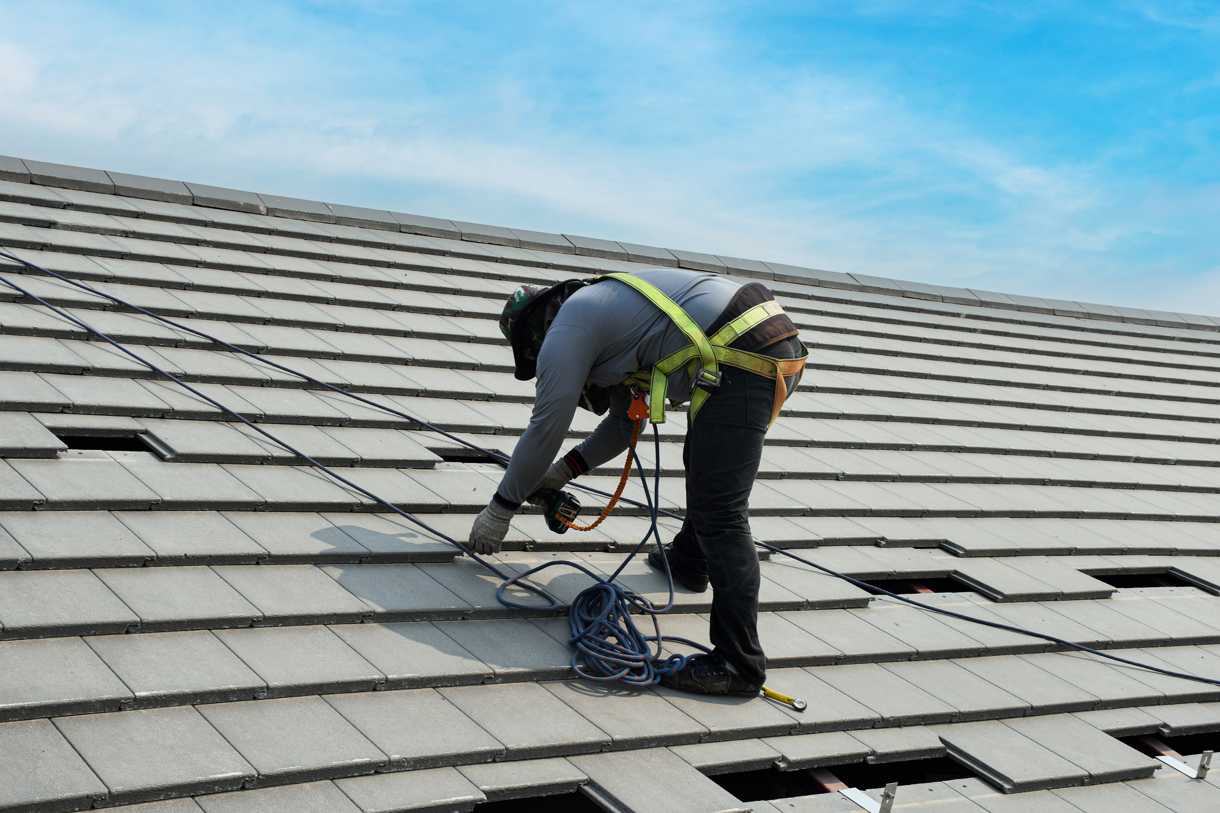 A construction worker wearing safety gear installs or repairs tiles on a sloped roof, using tools and secured by a harness and ropes, under a bright blue sky.