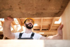 A construction worker wearing a yellow hard hat, safety glasses, and overalls stands under a wooden structure, looking up and holding onto beams at a construction site. Sunlight filters through the unfinished roof above.
