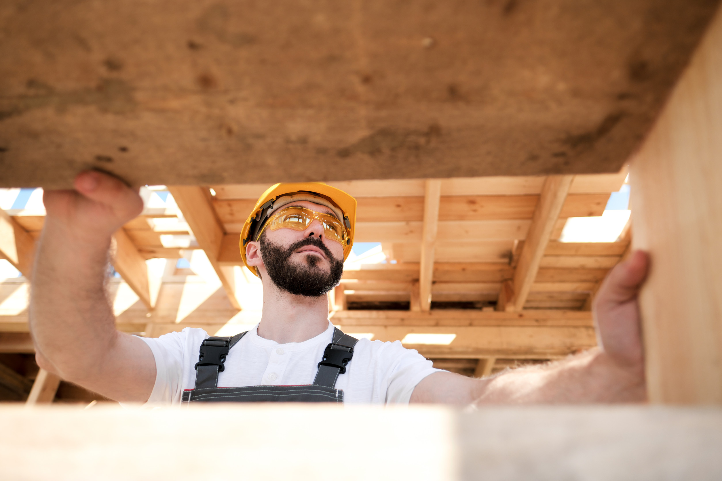 A construction worker wearing a yellow hard hat, safety glasses, and overalls stands under a wooden structure, looking up and holding onto beams at a construction site. Sunlight filters through the unfinished roof above.