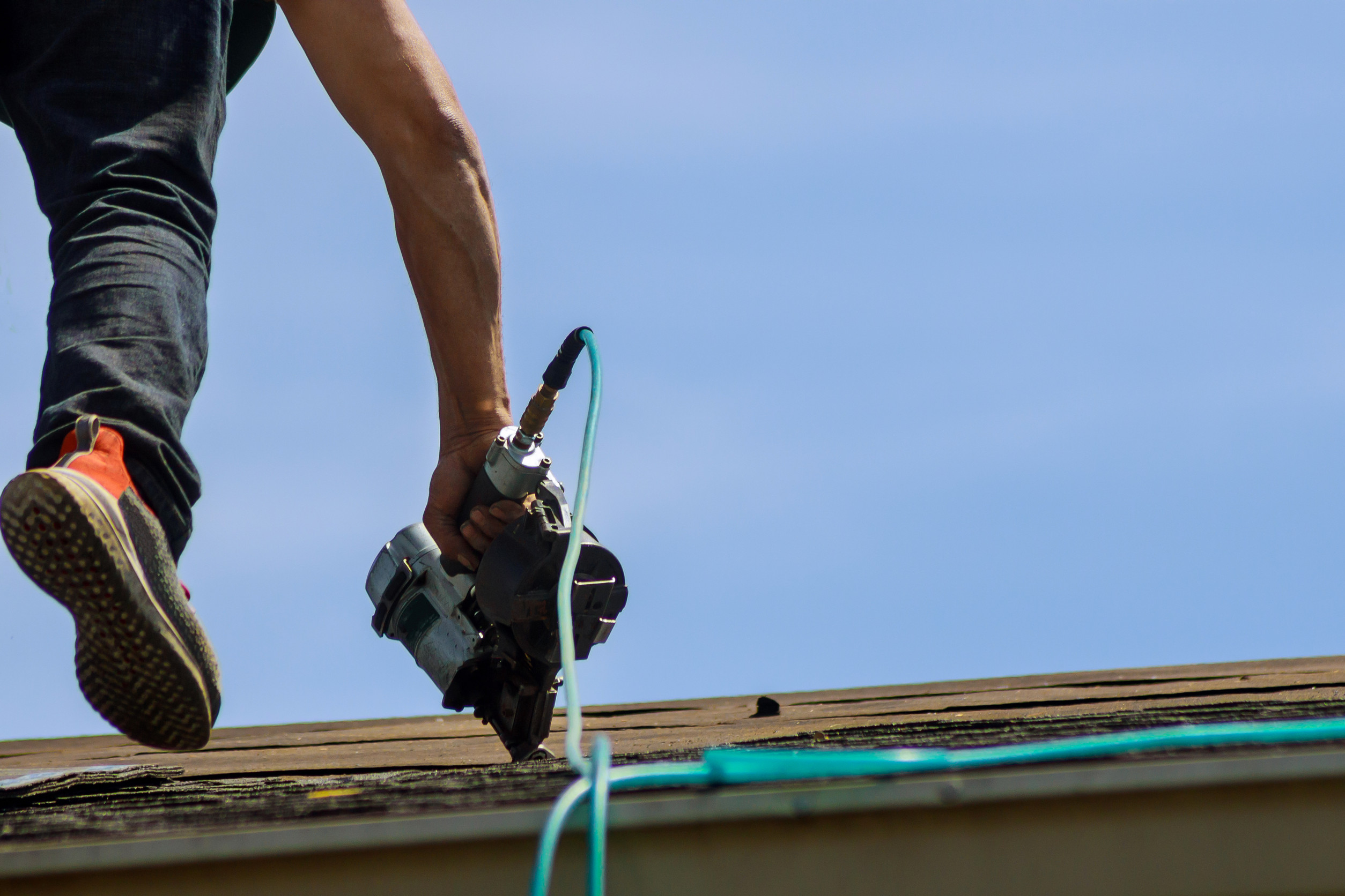A person on a roof holds a power tool, possibly a nail gun, while wearing orange-soled shoes. The scene is outdoors under a clear blue sky.