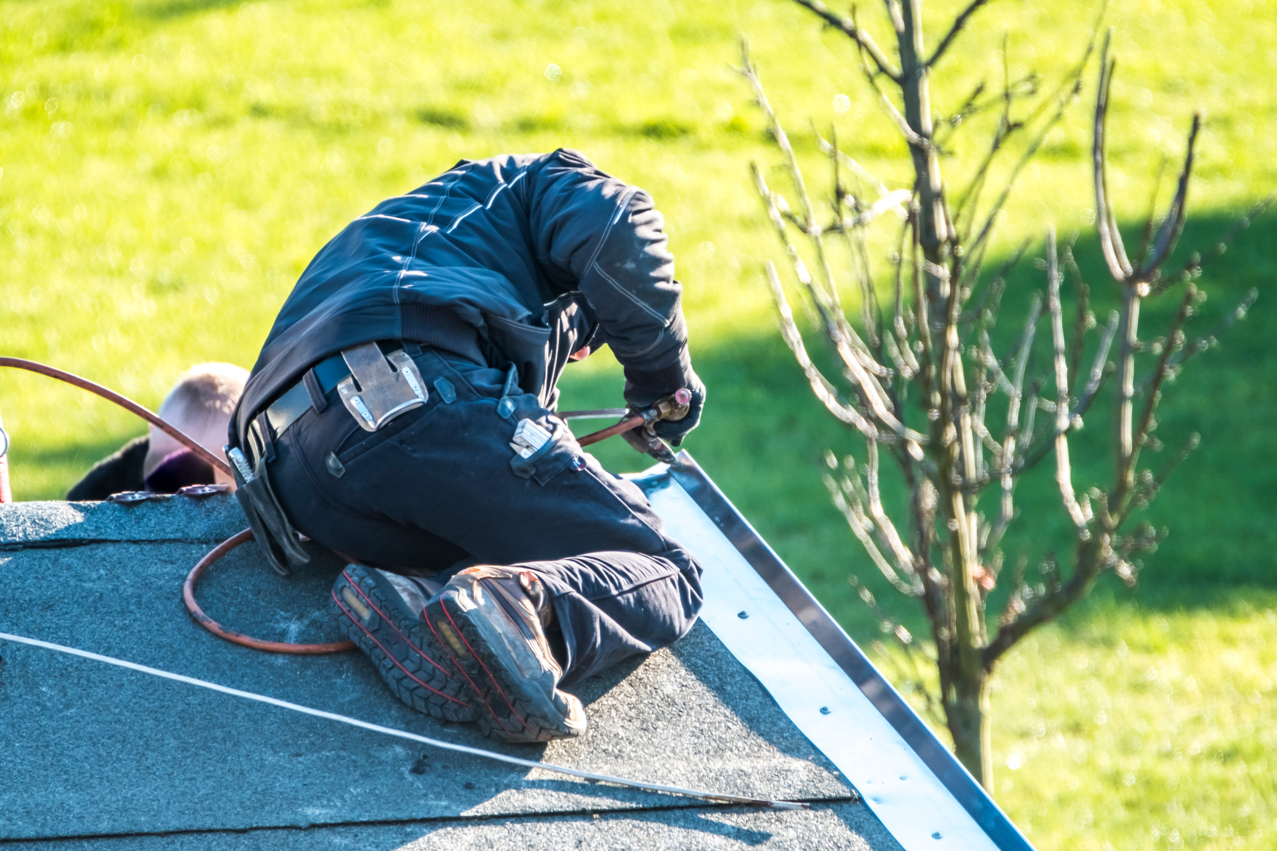 A worker in a black jacket and safety gear kneels on a roof, using tools to repair or install roofing material. A hose is attached to their tool, and a grassy area with a tree is visible below.