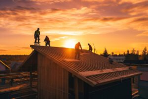 Construction workers install a roof on a wooden building at sunset, silhouetted against an orange sky with scattered clouds. The scene captures activity and teamwork on the rooftop.