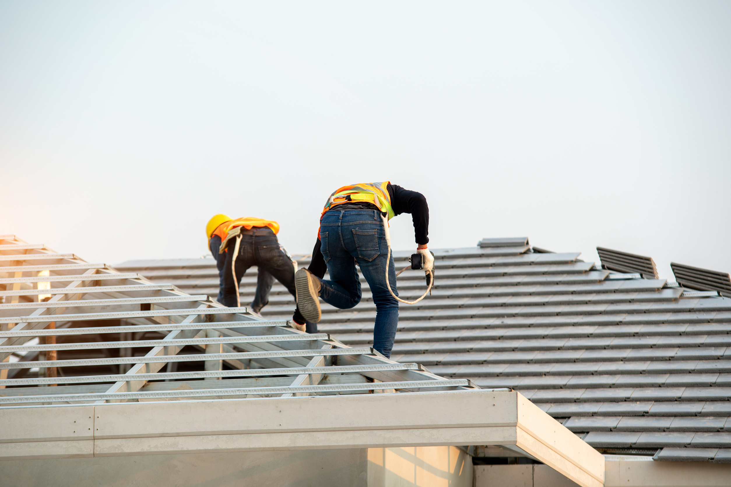 Two construction workers wearing safety helmets and vests are installing or inspecting metal roofing beams on a building in the Brainerd Lakes area. One worker is crouched, while the other climbs across the roof structure under clear skies.