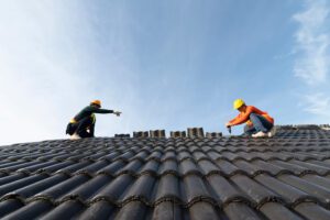 Two workers wearing safety gear and helmets are installing or inspecting tiles on a sloped roof under a clear blue sky. One worker is reaching toward the other across the rooftop.