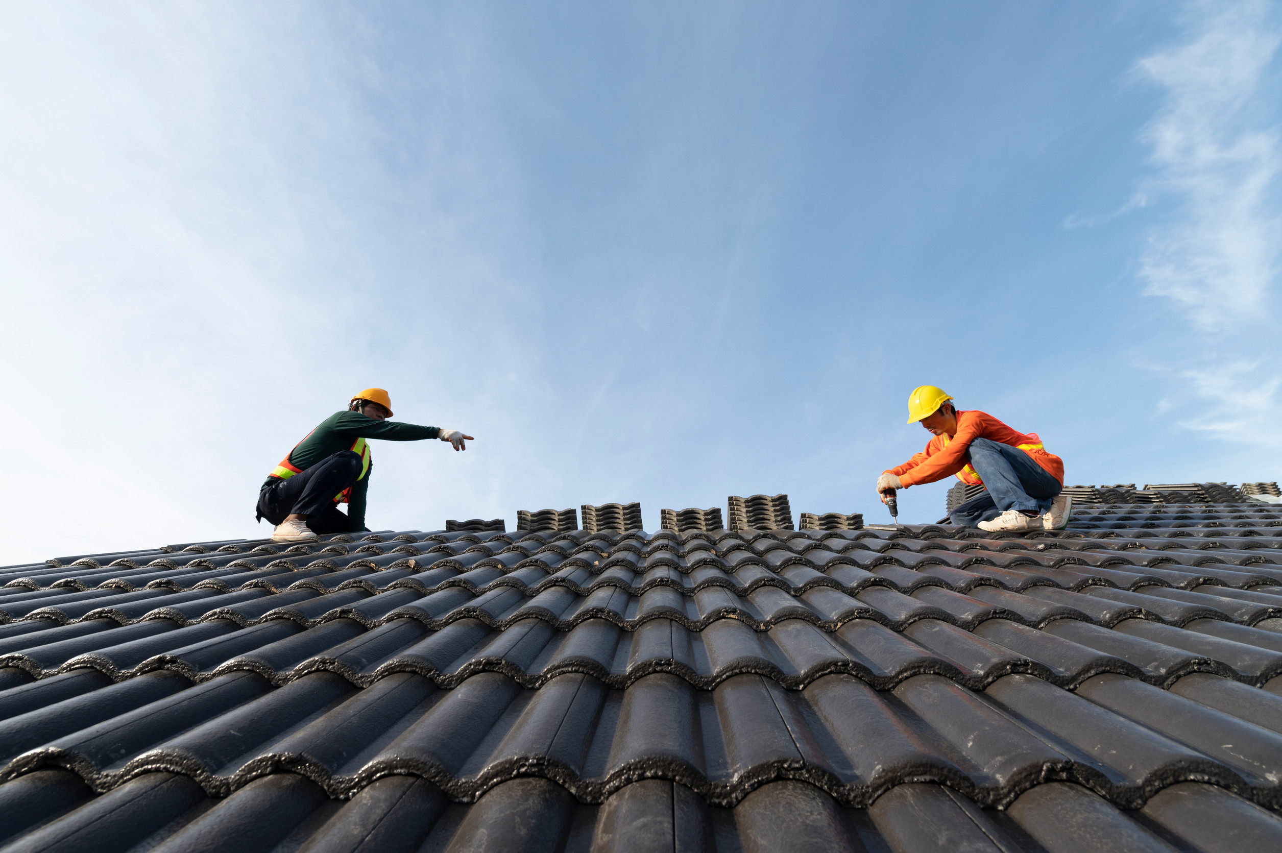 Two workers wearing safety gear and helmets are installing or inspecting tiles on a sloped roof under a clear blue sky. One worker is reaching toward the other across the rooftop.