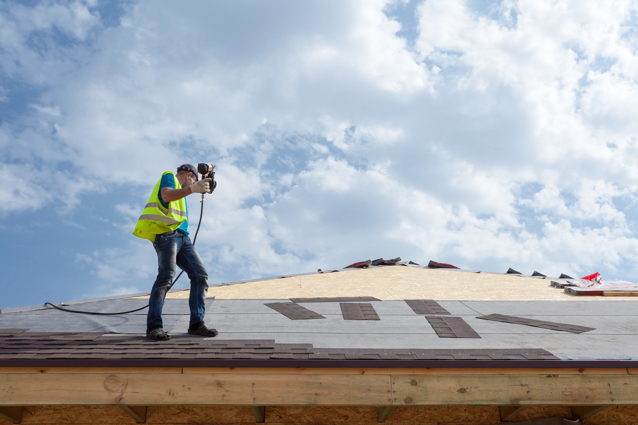 A construction worker in a yellow safety vest uses a nail gun to install roof shingles on a house under a partly cloudy sky. Roofing materials are scattered nearby on the roof.