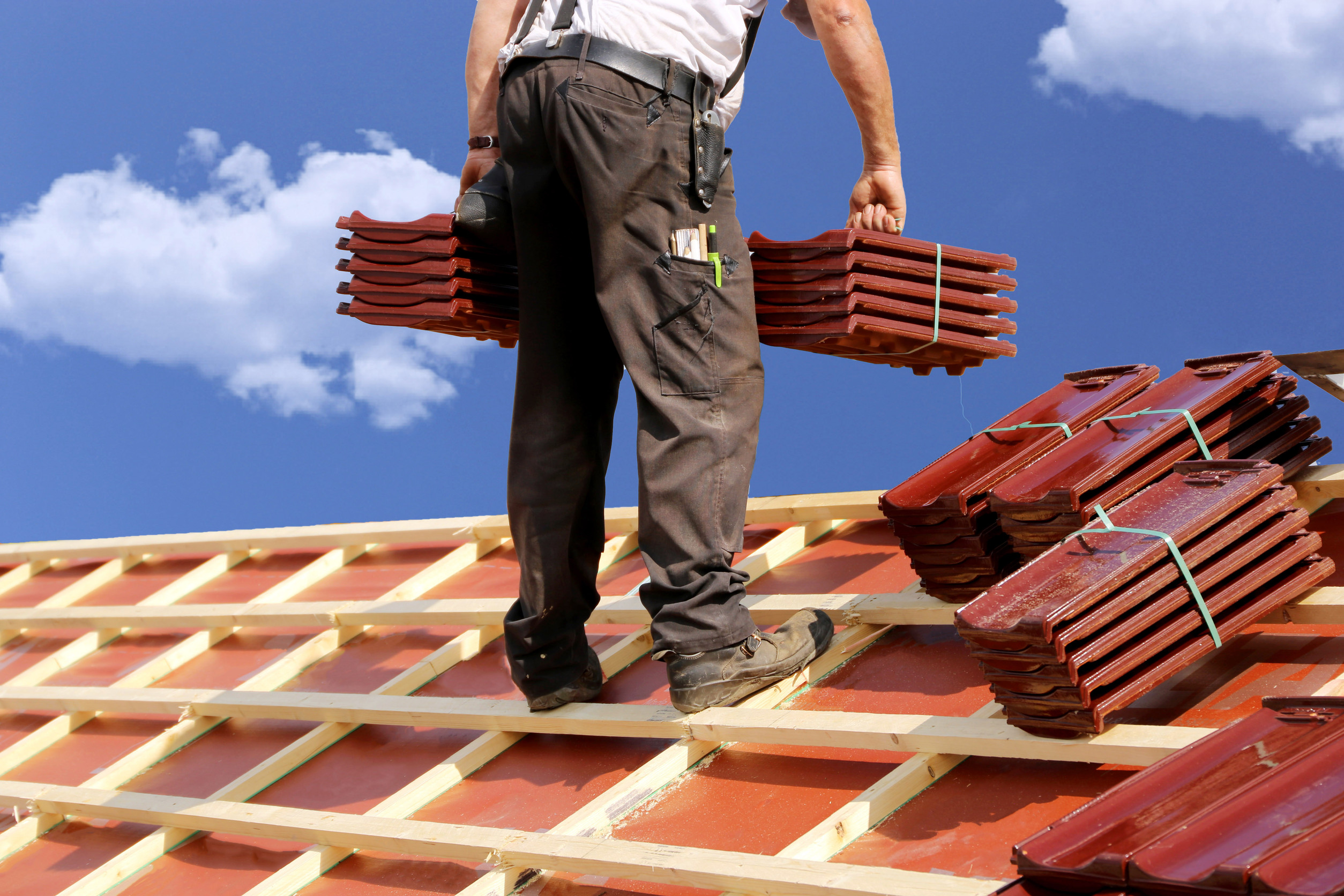 A worker in dark pants stands on a wooden roof frame, carrying stacks of red roof tiles under a blue sky with clouds. More roof tiles are stacked nearby on the roof—a scene showcasing expert roofing in action.